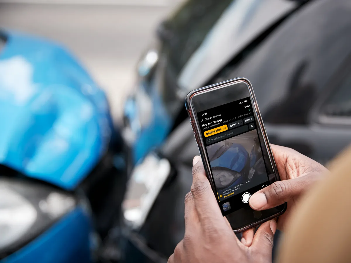 Two hands hold a phone with the EverStamp camera open by a blue car with a damaged front