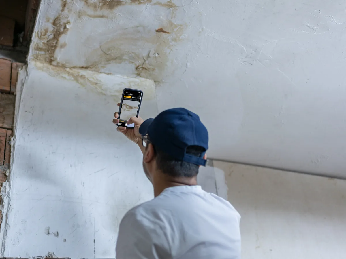A man in a cap photographs a leak in the ceiling with his phone; on the screen, the EverStamp camera